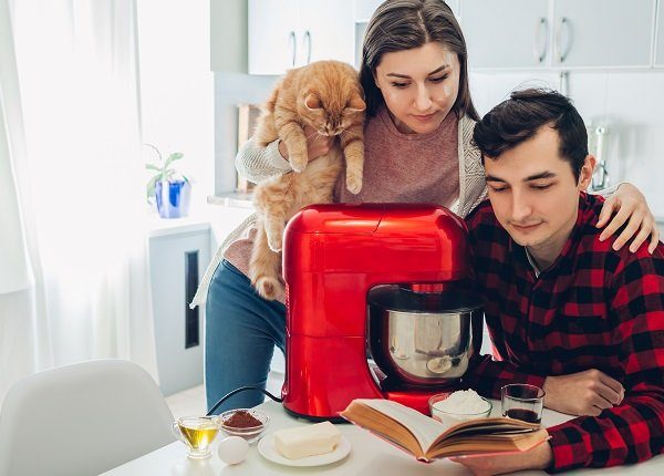 Young happy couple cooking on kitchen with food processor and holding cat. Woman and man reading recipe and hugging. Lifestyle