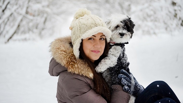 A young lady in winter gear holds a small dog with a jacket in front of a snowy forest background.