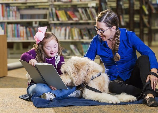 An 8 year old girl with down syndrome reading in the library, sitting next to a therapy dog and trainer, a mature woman in her 50s. The goldendoodle is trained as a reading assistance dog. The child is smiling as she holds the book up for the dog to read.