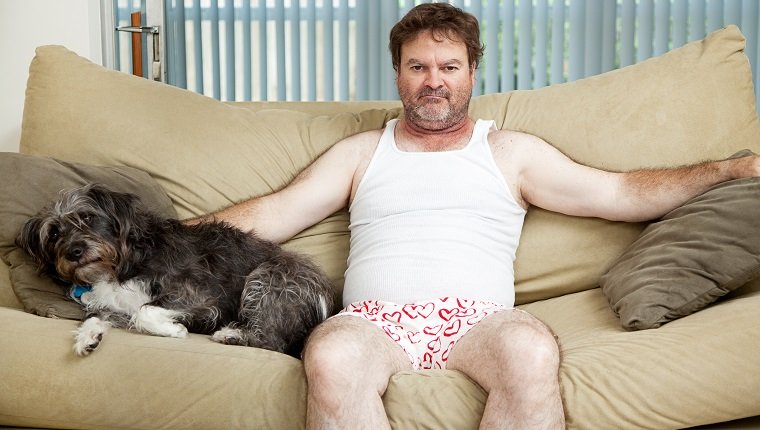 Entmutigter arbeitsloser Mann zu Hause in seiner Unterwäsche, mit seinem Hund auf der Couch sitzend.