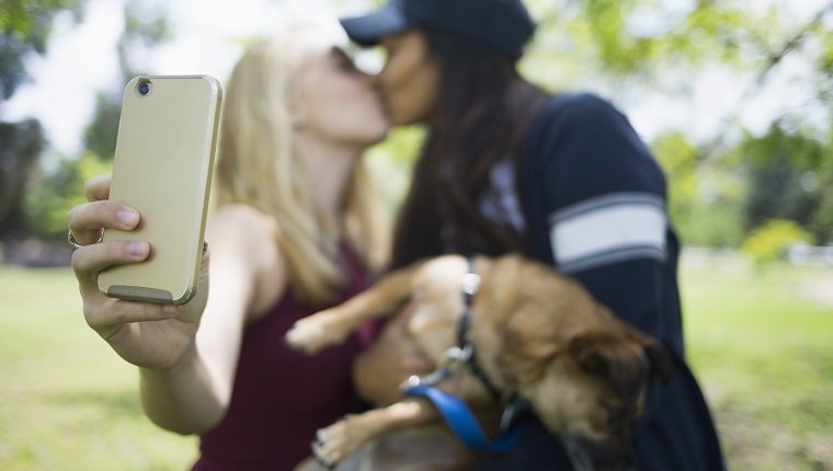 Frauen, die küssen, während sie selfie durch Smartphone mit Hund am Park nehmen