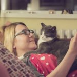 Beautiful young blonde woman is trying to read a book while her persistent cat is trying to sleep on her chest.
