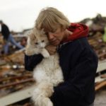 A woman holds her cat in front of a house that has been destroyed by a tornado.