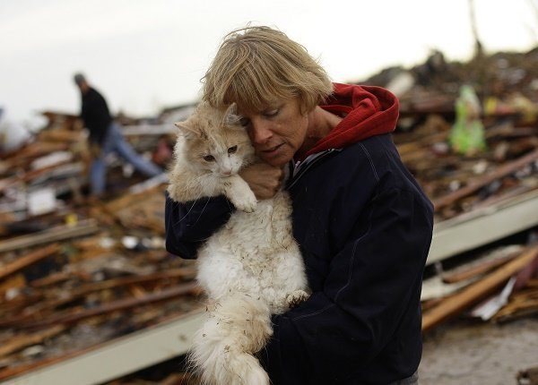 A woman holds her cat in front of a house that has been destroyed by a tornado.