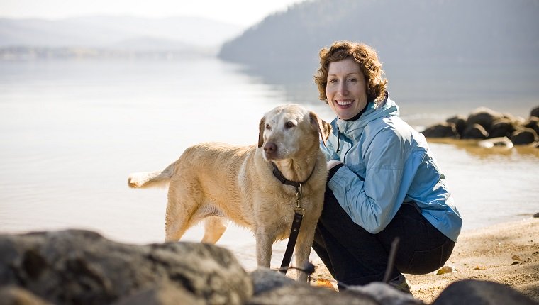 A smiling young woman with red hair and a blue jacket sits next to a golden labrador on the shores of Priest Lake. Priest Lake, Idaho.