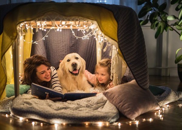 Young happy mother reading a book to her small girl while being in a tent at home with their dog.