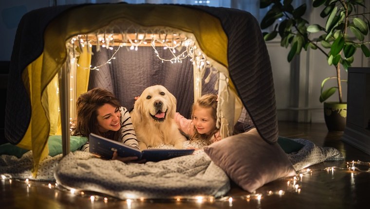 Young happy mother reading a book to her small girl while being in a tent at home with their dog.
