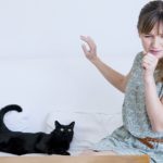 A woman sitting next to a black cat raises her hand to cover a sneeze.