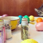 Frisky and mischievous cat reaching out to touch Easter eggs on a table. Little bowls filled with dye and colorful Easter eggs decorated in a rainbow of colors. Tiny jars of sparkling glitter scattered across the messy table. Childhood.