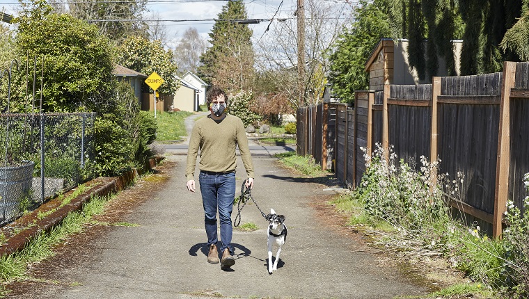 Porträt eines Mannes, der seinen Hund im Frühjahr eine Gasse entlangführt und eine hausgemachte Gesichtsmaske trägt.