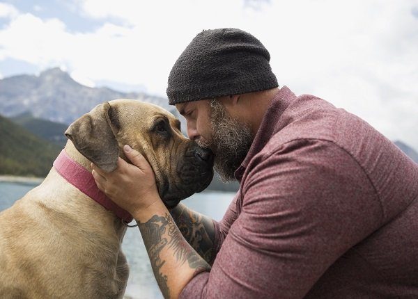 Affectionate bearded man with tattoos kissing dog at remote lakeside