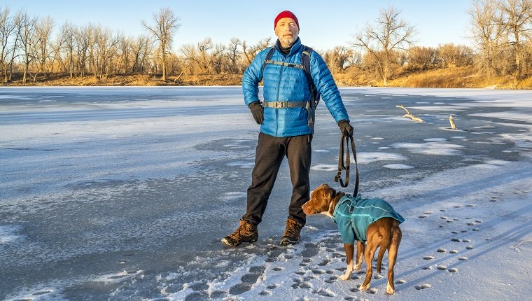 Ein älterer männlicher Wanderer mit einem Pitbull-Hund genießt den Sonnenuntergang an einem Ufer eines zugefrorenen Sees