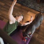 An adult woman does yoga and strength training exercises on a mat in her living room, her pet dog keeping her company and trying to play.