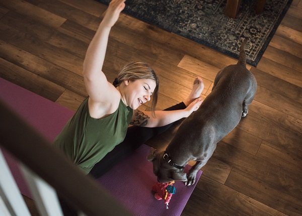 An adult woman does yoga and strength training exercises on a mat in her living room, her pet dog keeping her company and trying to play.