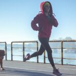 Young fit woman accompanied with her dog, running and listening to music on the riverbank in the city. The woman wears black running tights, yellow hat and red jacket. She has a smart phone mounted on her arm, with earphones connected to it. Her body is in motion, slightly above the ground. The copy space has been left. Shallow DOF.