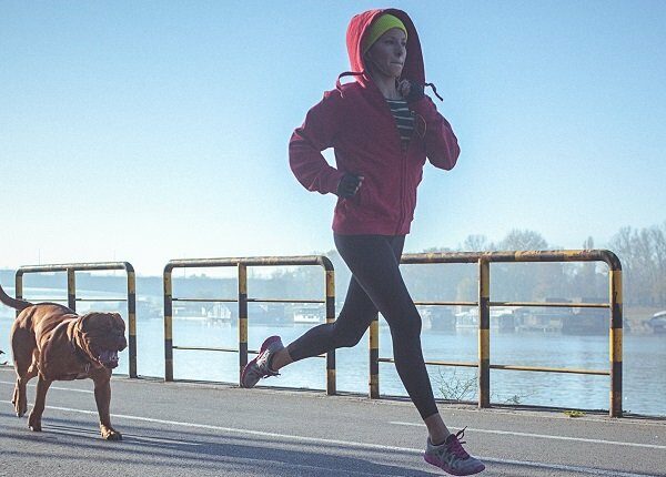 Young fit woman accompanied with her dog, running and listening to music on the riverbank in the city. The woman wears black running tights, yellow hat and red jacket. She has a smart phone mounted on her arm, with earphones connected to it. Her body is in motion, slightly above the ground. The copy space has been left. Shallow DOF.