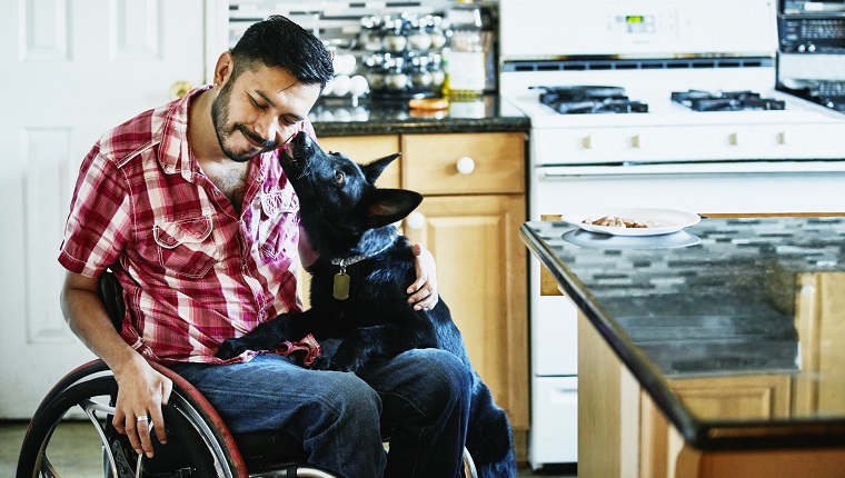 Lächelnder Mann im Rollstuhl, dessen Gesicht vom Hund geleckt wird, während er in der Küche rumhängt