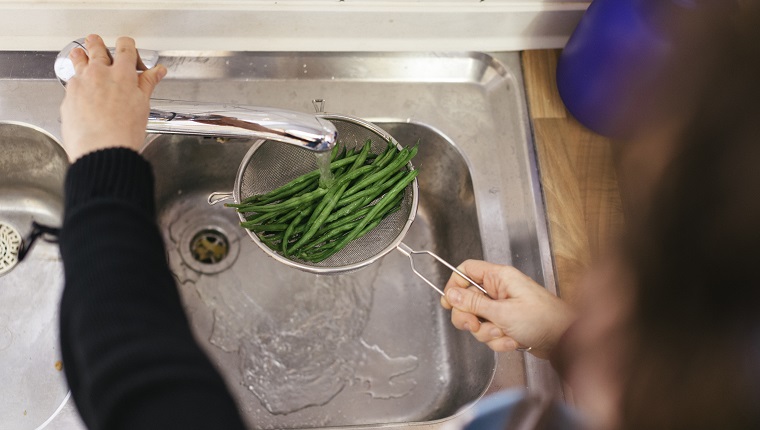 Schließen Sie oben von den grünen Bohnen, die mit Wasser in einem Küchenspülbecken gewaschen werden. Sie werden von einer Frau unter Wasser gehalten, die auf dem Foto nicht sichtbar ist.