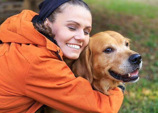 Smiling young woman kneeling and embracing a dog. Wears casual clothes, with eyes closed. Focus on foreground.