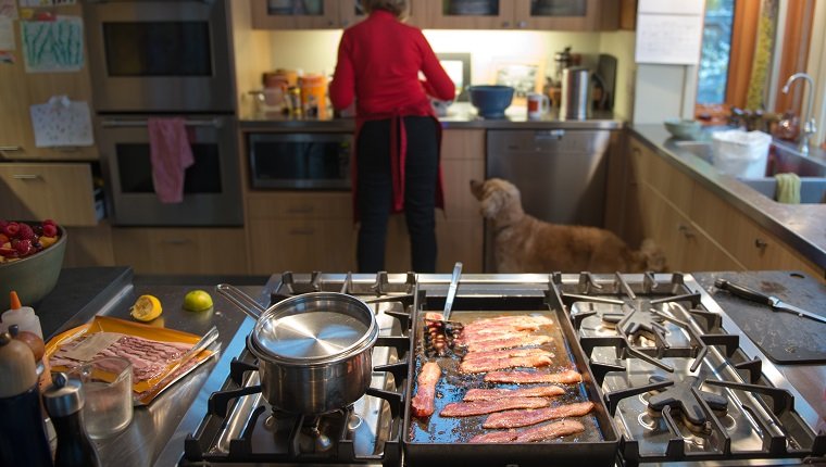 Großmutter bereitet in der Küche einen Brunch mit Speck und Eiern zu, während ihr Hund interessiert zusieht, wie sie die Zutaten mischt.