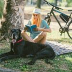 Mid adult woman enjoying the summer day with her dog, great dane, in the park. They are sitting on blanket and woman is reading book.